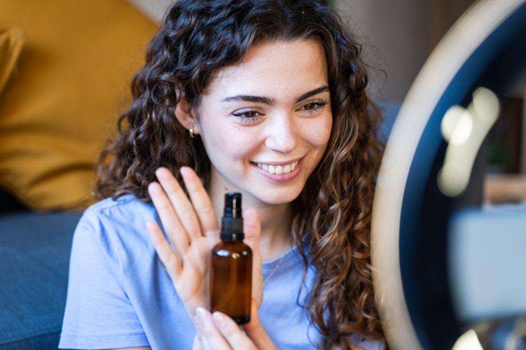 Curly-haired woman showing a skincare oil bottle while recording a video with a laptop and ring light